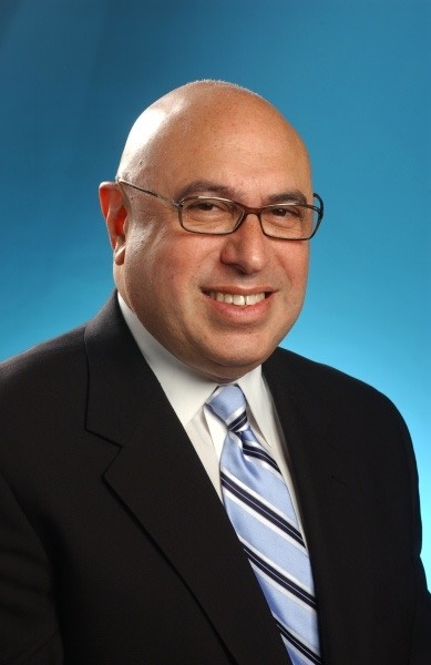 Dr. Mel J. Colón smiling in a dark suit and blue striped tie against a blue studio background