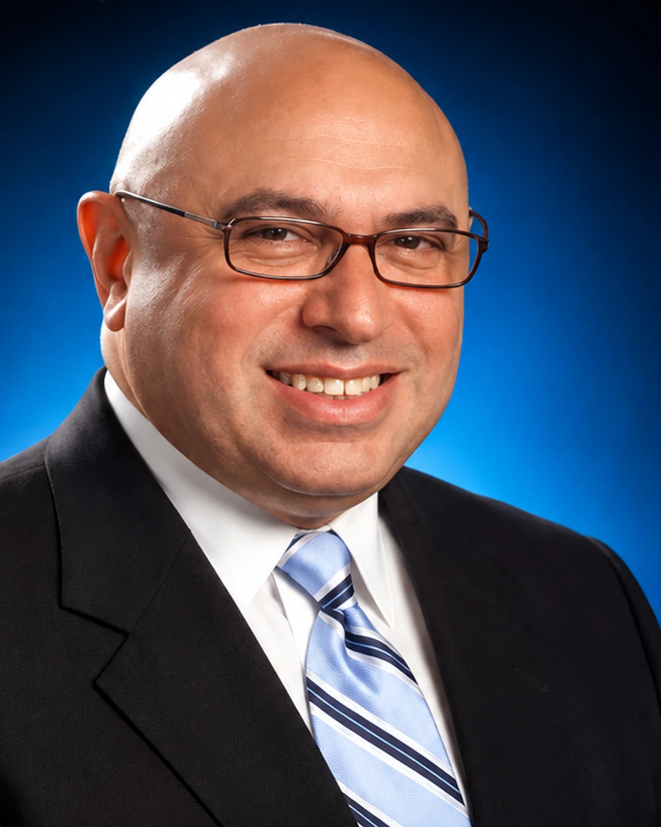 Dr. Mel J. Colón smiling in a dark suit and blue striped tie against a blue studio background